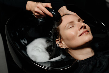 hairdresser washing client hair with water and foaming shampoo in black basin in beauty salon, massaging head, young beautiful caucasian woman getting hair washed, treatment and relax