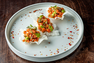 A gourmet dish on a pale plate featuring three crispy shells filled with a colorful mix of diced vegetables, garnished with microgreens and sprinkled with red chili flakes.