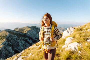 Young woman tourist stands on top of mountain, enjoys sunset on cliff. Traveler with backpack feels freedom, looks at mountain landscapes. Travel concept, freedom. Active lifestyle. © maxbelchenko