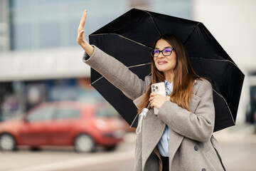 Young lady with umbrella calling a taxi on rainy weather on city street downtown.