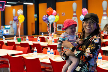 A bowling alley set up for children's birthday celebrations with birthday tables with balloons and water bottles and wall with drawn bowling pins. A smiling host mom with her daughter.