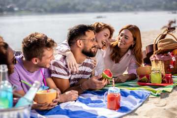 Group of five multiracial friends having picnic on the beach. They eat watermelon, they drink colorful cocktails and they have a guitar near them.