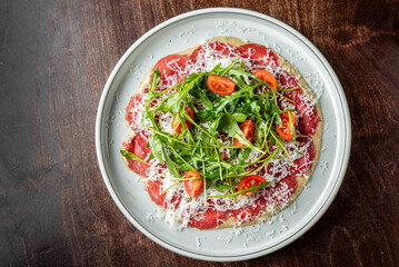 A beautifully plated raw beef carpaccio topped with fresh arugula and cherry tomatoes, garnished with grated cheese on a rustic wooden table.