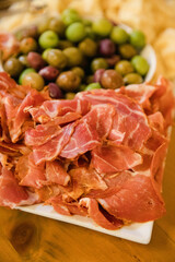 Slices of cured meat and bowl of olives are displayed on a wooden table at a casual gathering