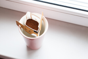 Drip bag in a ceramic cup on windowsill. Instant freshly brewed alternative filter drink in mug. Easy brew black coffee method for home, travel or office. cozy trendy lifestyle