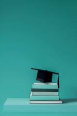 A black graduation cap placed on a neatly stacked pile of books against a teal background. A symbolic representation of knowledge, success, and academic achievement.
