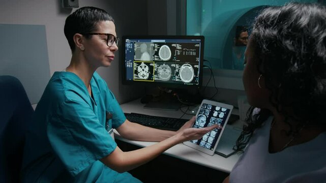 Medium shot of smiling middle-aged Caucasian female radiologist and African American patient discussing brain MRI scan on digital tablet, doctor sharing news of remission from tumor treatment