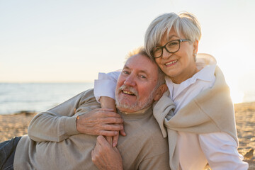 Senior mature couple sitting on beach hugging enjoying outdoor recreation. Old husband wife embracing with tenderness love enjoying sweet bonding. Grandmother grandfather together. Family moment love