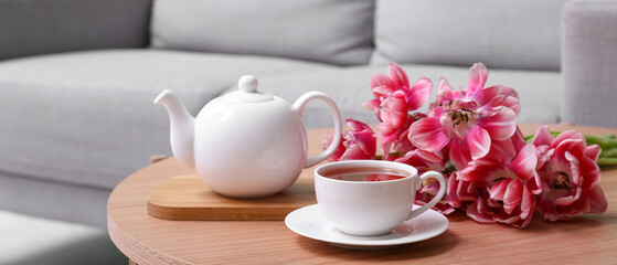 White teapot and cup of hot tea with pink tulip flowers on table in living room