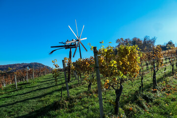 Rustic Klopotec windmill stands sentinel over a vineyard in Ratsch an der Weinstrasse, Styria, Austria. Wooden blades contrasting against clear blue sky. Rolling hills in distance. Styrian wine region