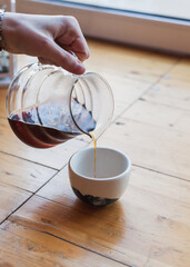 Barista pouring filter coffee from kettle to a ceramic white cup. female or man hand holding glass teapot server with black pour over coffee. alternative brewing, specialty v60. background light wood