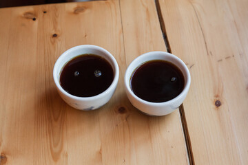 Young caucasian freelancer woman hold two white cup's with black filter coffee. Barista on cafeteria giving mug to customer top view. alternative brewing of specialty coffee. background light wood