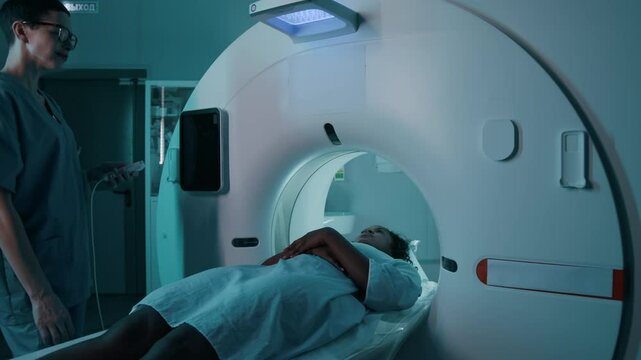 Medium shot of middle-aged Caucasian female radiologist pressing control button, and African American patient sliding through MRI scanner during brain tomography procedure to detect tumor