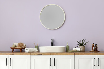 Obraz premium Mirror, chest of drawers with sink and bath supplies near lilac wall in interior of bathroom