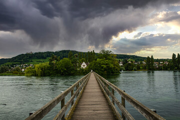 Dramatic clouds loom over a wooden bridge extending into calm waters, creating a serene yet...