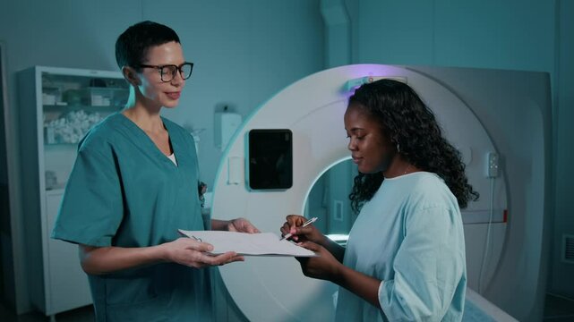 Medium shot of young African American woman in disposable gown sitting on sliding table by MRI scanner, signing medical consent form offered by doctor on clipboard before beginning procedure