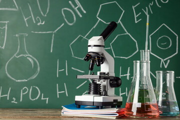 Modern microscope and flasks on wooden table near chalkboard with chemical formulas