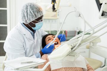 African-american man orthodontist using tools and doing oral care inspection on Asian woman patient...