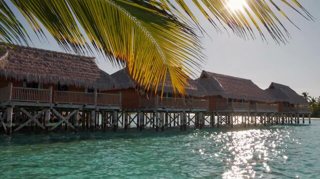 Tropical island with thatched-roof huts on stilts over the water, surrounded by turquoise lagoons and coral reefs.