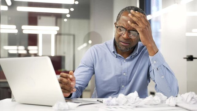 Exhausted african american businessman is bored sitting at workplace at desk in business office. Overworked black man is tired of doing paper work. Frustrated confused entrepreneur has depression
