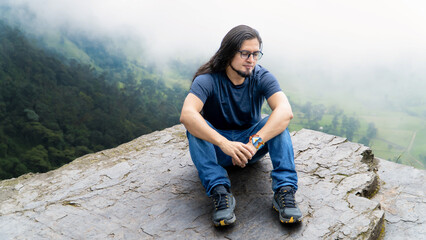 Naklejka premium Smiling young man sits on a rocky cliff with a misty green valley in the background. Peaceful outdoor adventure in nature