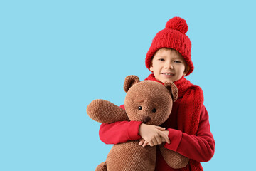 Cute fashionable boy in warm red sweater, scarf and winter hat with teddy bear on blue background
