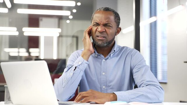 African american businessman suffers from ear pain sitting at workplace in office. Upset male touches his ear with his fingers. He has furuncle, shingles, eczema, ear diseases or otitis media. Earache