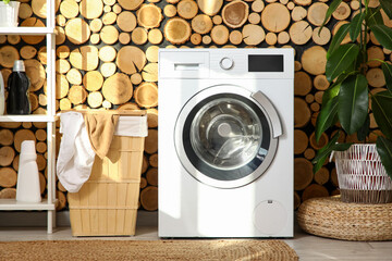 Modern washing machine with laundry basket and ficus houseplant near wooden wall in bathroom