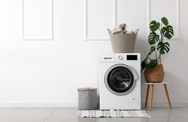 Modern washing machine with laundry basket near white wall in bathroom