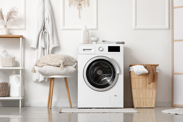 Modern washing machine with laundry basket and pillows on stool near white wall in bathroom