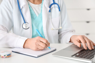 Mature female doctor with laptop writing on clipboard in clinic, closeup