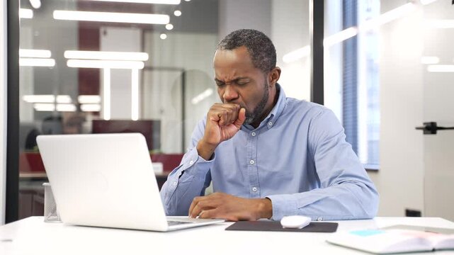 African american businessman coughing while working on a laptop sitting at desk in business office. Sick with a cold or flu black man worker feels bad, holds his hand to his chest. He has bronchitis