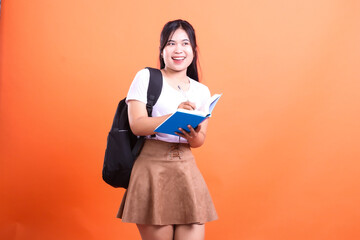A young woman with a black backpack is writing in a blue book while looking sideways against orange background