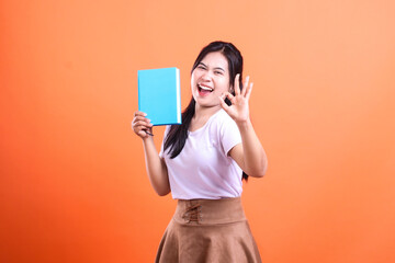 Naklejka premium A smiling woman in a white t-shirt holds a blue book, with her hand making the okay sign, isolated on orange background