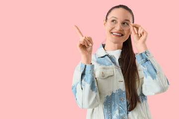 Beautiful young happy woman in new eyeglasses pointing at something on pink background