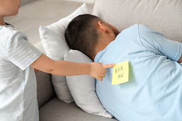 Little son attaching paper sheet with text KICK ME to his father's back at home. April fool's day...