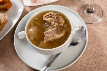 Freshly prepared hearty lunch awaits on table - fragrant cabbage soup with potato, carrot and pork rib. Dish is complemented with plate of bacon and bread.