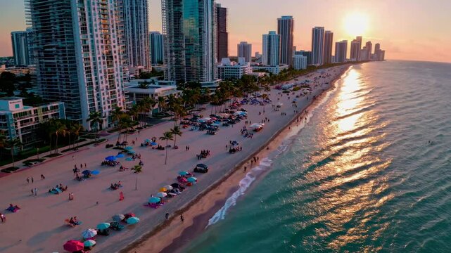 Aerial video captures a vibrant beach cityscape at sunset, with high-rise buildings and palm trees framing the golden horizon and shoreline.