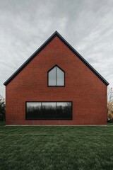 Obraz premium Modern red brick house with unique roof design and large windows located in a serene environment surrounded by green grass on a cloudy day