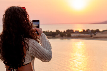 A woman holding a smartphone taking pictures outside during sunrise or sunset. Travelling and vacation concept.
