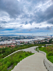 A scenic view showcases a meandering stairway leading through lush greenery, guiding the eye to a sprawling cityscape under a cloudy sky. the city, bordered by a river, is vibrant with urban buildings