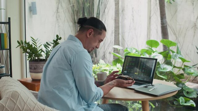 Medium long shot of male multi-ethnic freelancer holding small python while sitting in front of laptop, analyzing charts at cozy home office