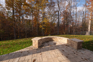 A serene backyard with a curved stone wall and patio pavers, surrounded by bare trees, autumn foliage, and scattered leaves.