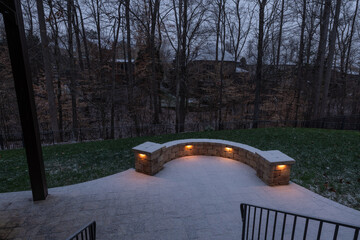 A peaceful backyard patio covered in fresh snow, featuring a curved stone wall with glowing lights. Black railings and bare trees enhance the serene winter ambiance at dusk.