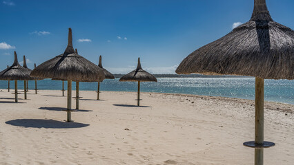 Rows of straw umbrellas on a sandy beach. Round shadows on white sand. There are no people. Highlights, sparks on the surface of a calm turquoise ocean. Blue sky, clouds. Mauritius. The resort hotel.