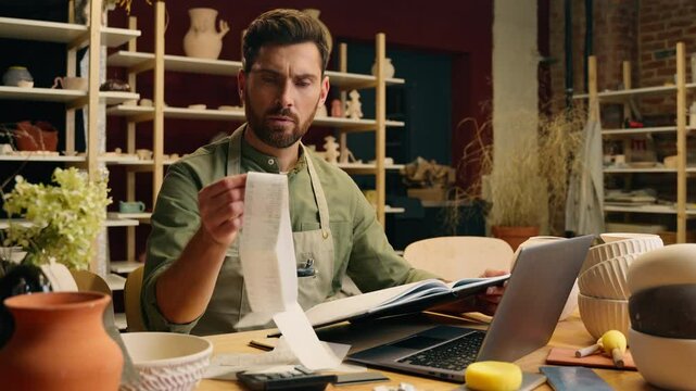 Caucasian man businessman male guy small business owner entrepreneur craftsman potter writing notebook examining check receipt working laptop calculate accounting finance budget inside pottery store