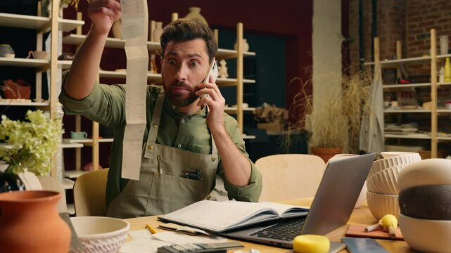 Shocked nervous worried Caucasian man businessman male small business owner entrepreneur potter examine large receipt working laptop accounting talking mobile phone call conflict inside pottery store
