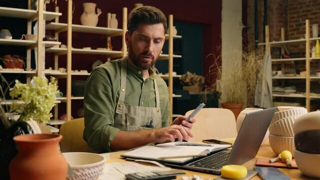 Caucasian man guy businessman male small business owner entrepreneur potter counting money dollars banknotes looking bill receipt shock high cost expense finance management laptop inside pottery store