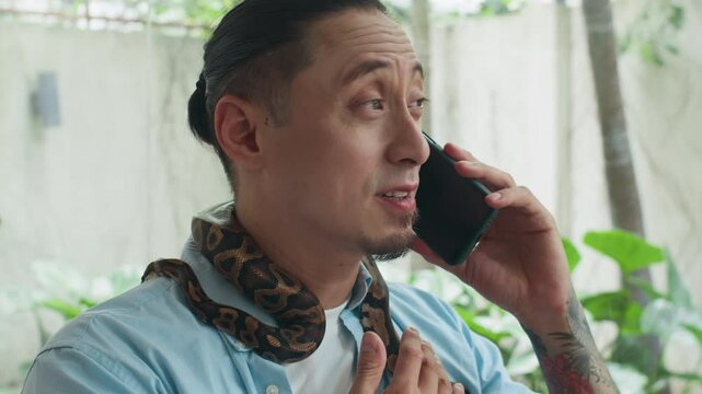 Tilt up shot of tattooed man calling on smartphone while interacting with pet python wrapping around his neck at home