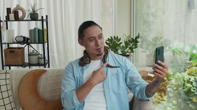 Medium shot of relaxed multicultural man in casual shirt talking on video call in front of smartphone while petting python resting on his neck at home